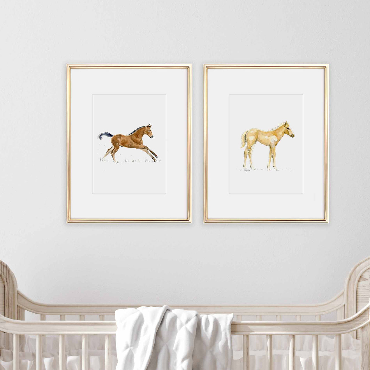 two framed baby horse prints, one is a brown foal running and one is a palomino foal standing, they are showin in thin gold metal frames with 2 inch white mats, and are hanging above a baby's white crib