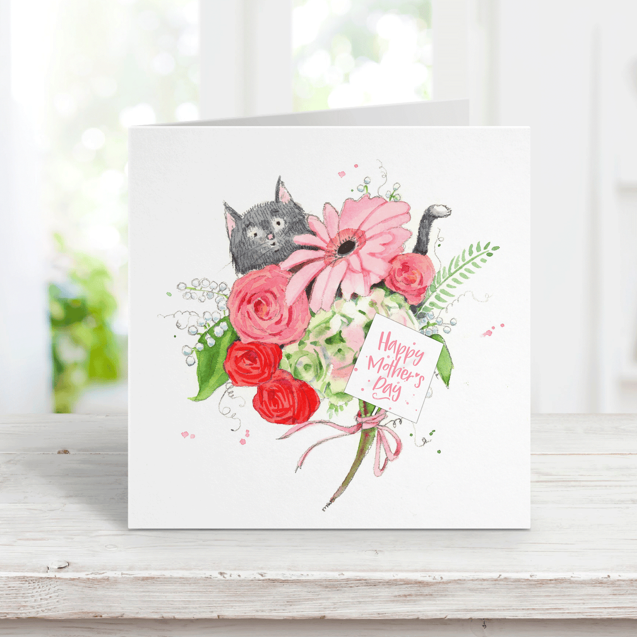 watercolor mothers day card of a whimsical gray cat peeking out from behind a bouquet of pink and red flowers with a little tag that says Happy Mothers Day