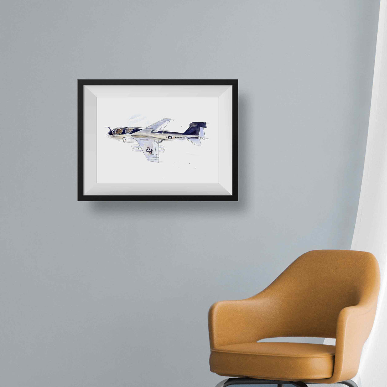 An EA-6B Prowler Airplane watercolor print framed with a white mat and black frame on a gray wall with a modern style tan chair