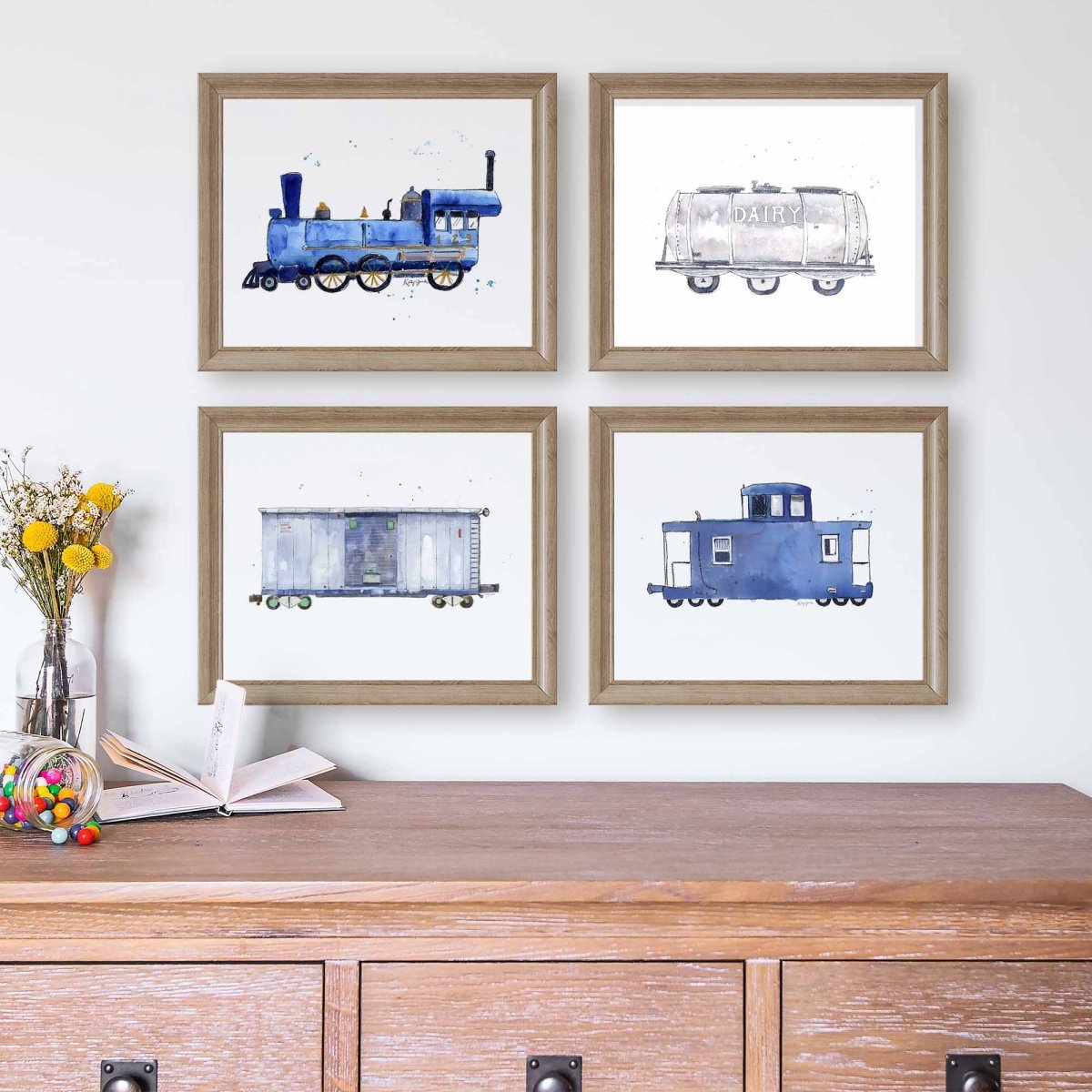 Four framed watercolor train illustrations arranged in a 2x2 grid on a white wall above a wooden cabinet. The prints show a blue steam locomotive, a dairy tank car, a gray boxcar, and a blue caboose, each in a light wood frame.