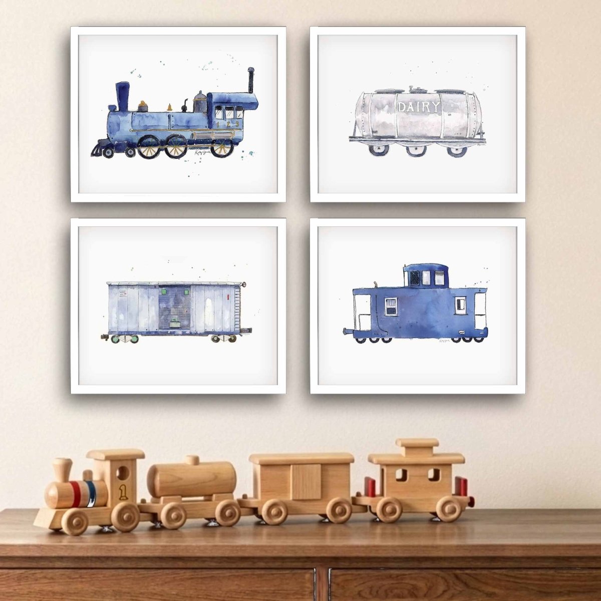 Four framed watercolor train illustrations arranged in a 2x2 grid on a beige wall above a wooden surface with a wooden toy train. The prints show a blue steam locomotive, a dairy tanker car, a blue cargo car, and a blue caboose.