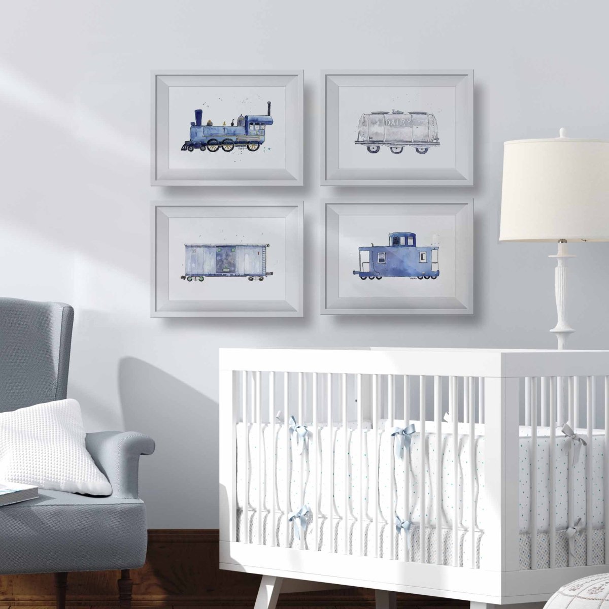 A styled nursery scene with four framed watercolor train illustrations arranged in a 2x2 grid on a light gray wall above a white crib. A gray armchair, white pillow, and white table lamp are visible in the room.